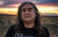 Portrait of artist Raven Chacon wearing a black tee shirt with white graphic, standing in an arid field with the sun low on the horizon.